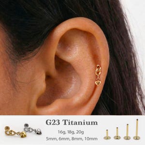 May include: A gold-colored heart charm earring with a chain link design. The text G23 Titanium, 16g, 18g, 20g, and 5mm, 6mm, 8mm, 10mm is visible. The earring is shown inserted into an ear cartilage piercing, alongside silver and gold options.