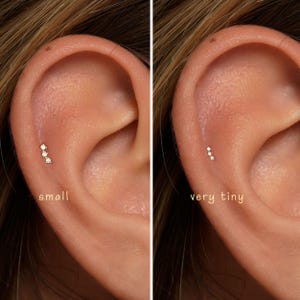 May include: A gold-toned ear cartilage stud featuring three small, clear, round-cut stones arranged in a vertical line. The image displays two variations of the stud, labeled small and very tiny, positioned on the upper ear cartilage.