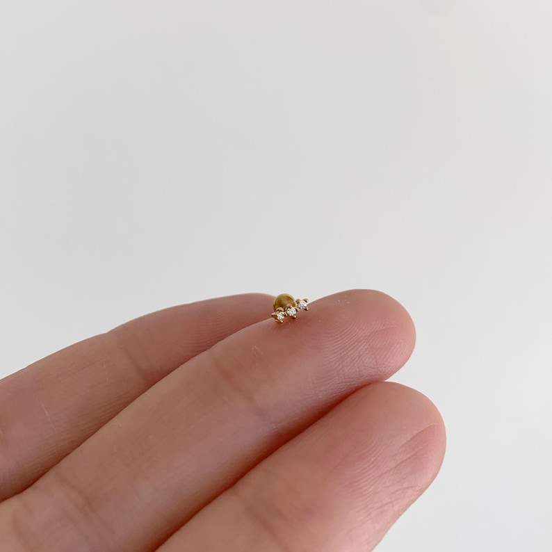 Pu&ograve; includere: Un piccolo orecchino a lobo color oro con un gruppo di tre minuscole pietre trasparenti a taglio rotondo disposte in fila orizzontale. L'orecchino presenta una finitura in metallo lucido e una chiusura sicura per il foro all'orecchio.