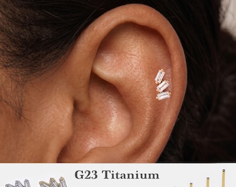 May include: A G23 titanium ear piercing stud featuring three clear baguette-cut stones arranged in a cluster. The text G23 Titanium, 16g, 18g, 20g, and 5mm, 6mm, 8mm, 10mm is visible. The jewelry is shown in silver and gold tones on a flat surface.