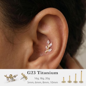 May include: A gold-tone G23 titanium ear piercing stud featuring a branch design with marquise-cut clear stones. The text G23 Titanium, 16g, 18g, 20g, and 5mm, 6mm, 8mm, 10mm is visible, along with four gold-tone flat-back posts of varying lengths.