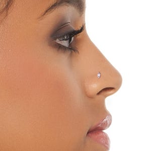 Pode incluir: Um close-up de um rosto, destacando um piercing no nariz. O piercing &eacute; um pequeno brinco oval com uma pedra clara e brilhante. O tom de pele &eacute; castanho m&eacute;dio quente, contra um fundo branco.