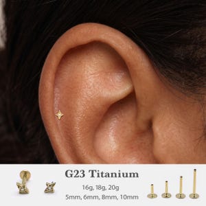 May include: A gold-coloured G23 titanium flat back stud earring featuring a four-pointed star design with a central clear stone. The text G23 Titanium, 16g, 18g, 20g, and 5mm, 6mm, 8mm, 10mm is visible alongside various post lengths and styles.
