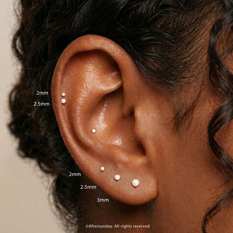 Peut inclure: Un ensemble de petites boucles d'oreilles &agrave; clous rondes et blanches port&eacute;es dans plusieurs piercings &agrave; l'oreille. Les clous sont &eacute;tiquet&eacute;s avec les tailles 2mm, 2,5mm et 3mm. Le bijou pr&eacute;sente un design circulaire simple avec une finition lisse.