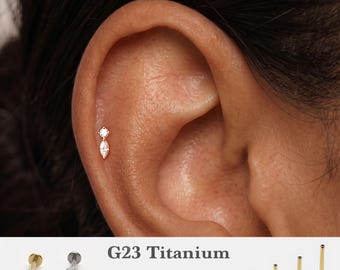Può includere: Un orecchino in titanio G23 con una piccola pietra rotonda trasparente sopra una pietra trasparente taglio marquise. Il testo recita G23 Titanium, 16g, 18g, 20g e 5mm, 6mm, 8mm, 10mm. L'immagine mostra borchie color oro e argento e quattro perni.