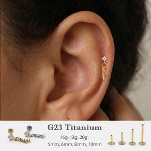May include: A gold-tone G23 titanium flat back stud earring featuring a small clear gemstone and a delicate dangling chain. The set includes various post lengths of 5mm, 6mm, 8mm, and 10mm, with gauge options of 16g, 18g, and 20g for the stud.