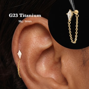 May include: A gold-tone G23 titanium earring featuring a kite-shaped clear gemstone stud connected to a delicate dangling chain. The jewelry is labeled 18g / 6mm. The design includes a flat back post, visible in the circular inset detail.