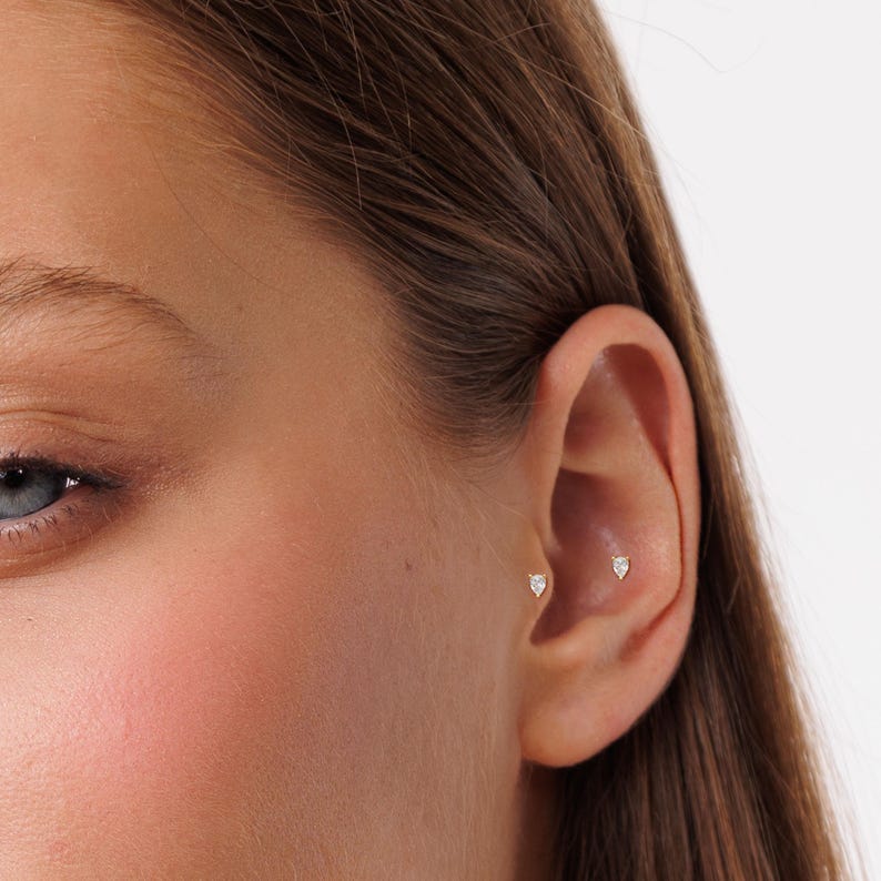 K&ouml;nnte beinhalten: Nahaufnahme eines Ohrs mit zwei kleinen Diamant-Ohrsteckern. Die Ohrringe sind in Gold gefasst und werden im Helix-Piercing getragen.