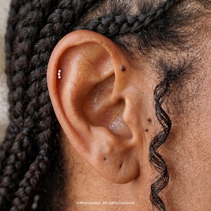 Pu&ograve; includere: Un piccolo orecchino da cartilagine color oro con tre minuscole pietre trasparenti allineate verticalmente. L'orecchino &egrave; posizionato sul bordo superiore esterno dell'orecchio, vicino a capelli scuri intrecciati e alcuni piccoli nei scuri.