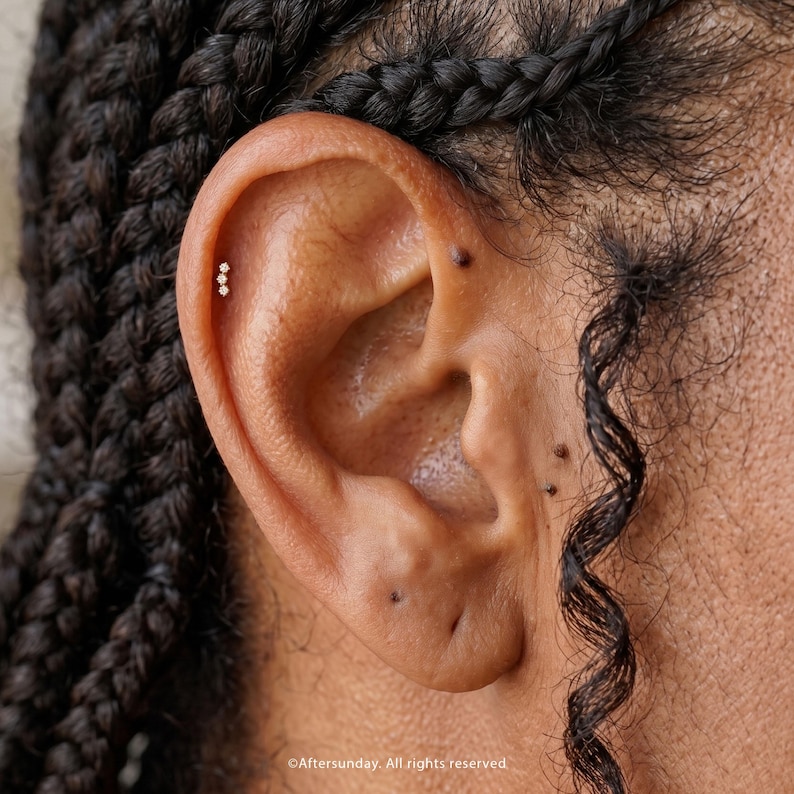 Pu&ograve; includere: Un piccolo orecchino da cartilagine color oro con tre minuscole pietre trasparenti allineate verticalmente. L'orecchino &egrave; posizionato sul bordo superiore esterno dell'orecchio, vicino a capelli scuri intrecciati e alcuni piccoli nei scuri.