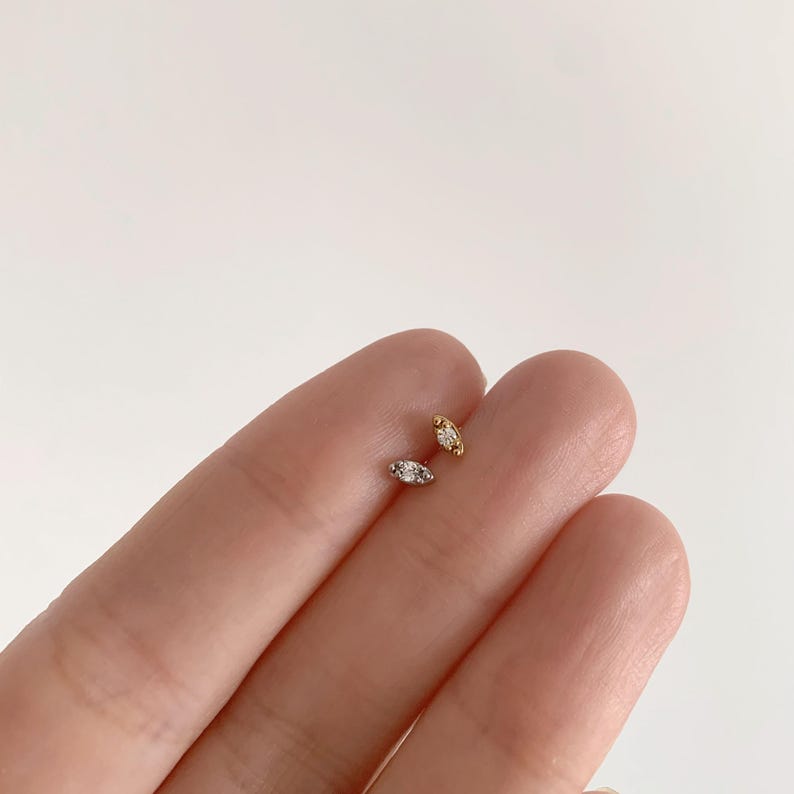 Pode incluir: Dois brincos pequenos, em forma oval, com uma pedra central transparente. Um brinco &eacute; dourado, o outro &eacute; prateado. Os brincos s&atilde;o exibidos sobre um fundo claro.
