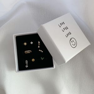 Op de afbeelding: Een wit vierkant sieradendoosje met de tekst LOVE LOVE LOVE en een smiley-icoon, met acht verschillende oorbellen waaronder ster-, bloem-, hart- en bladvormen, op een zwarte fluwelen inzetstuk.