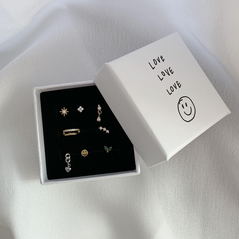 Op de afbeelding: Een wit vierkant sieradendoosje met de tekst LOVE LOVE LOVE en een smiley-icoon, met acht verschillende oorbellen waaronder ster-, bloem-, hart- en bladvormen, op een zwarte fluwelen inzetstuk.