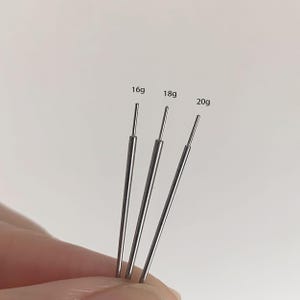 Può includere: Tre aghi argentati di diversi calibri, etichettati 16g, 18g e 20g. Gli aghi sono tenuti su uno sfondo bianco. Gli aghi sono usati per il piercing.