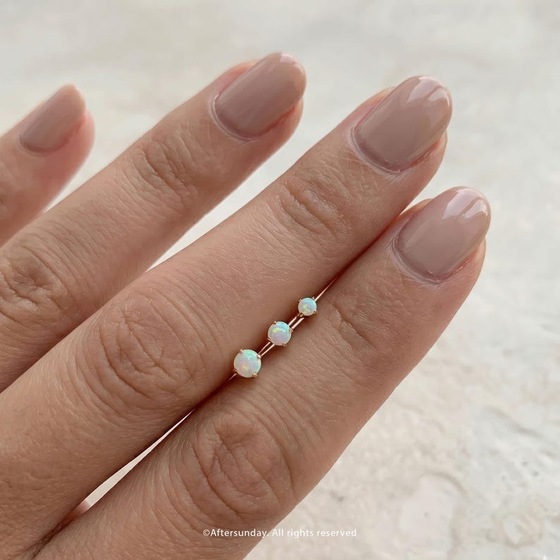 Peut inclure: Une bague d&eacute;licate en or orn&eacute;e de trois pierres d'opale rondes et iris&eacute;es dispos&eacute;es en ligne. Les pierres varient l&eacute;g&egrave;rement en taille, la plus grande opale &eacute;tant plac&eacute;e &agrave; une extr&eacute;mit&eacute; et la plus petite &agrave; l'autre, sur un anneau en or fin.