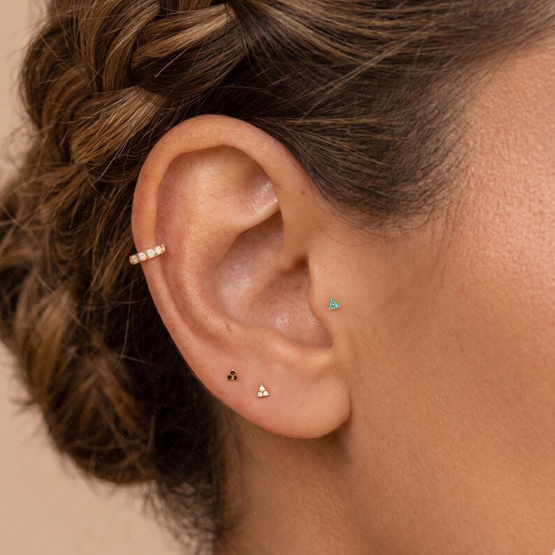 Op de afbeelding: Een gouden oormanchet met een rij kleine heldere stenen, een piepkleine groene driehoekige knop in de tragus en twee kleine driehoekige knoppen in de oorlel, &eacute;&eacute;n met zwarte stenen en &eacute;&eacute;n met heldere stenen, alles in gepolijst goud.