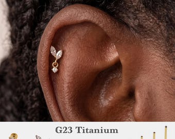 Può includere: Un orecchino da cartilagine in titanio G23 color oro con due pietre chiare taglio marquise e una pietra rotonda pendente da un piccolo anello. Il testo G23 Titanium, 16g, 18g, 20g e 5mm, 6mm, 8mm, 10mm è stampato sotto i gioielli.