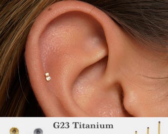 May include: A close-up of a person's ear with a small gold stud earring with two clear gemstones. The earring is made of G23 titanium and is available in sizes 5mm, 6mm, 8mm, and 10mm. The text "G23 Titanium" and "16g, 18g, 20g" is visible below the image.