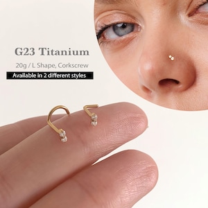 以下が含まれることがあります： ゴールドカラーのG23チタンノーズリング。1つはコルクスクリュー型、もう1つはL字型です。どちらも小さな透明な宝石が2つ付いています。テキストには「G23 Titanium 20g / L Shape, Corkscrew Available in 2 different styles」と書かれています。