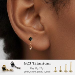 May include: A gold-colored earring with a black square gem and a dangling charm with a small white gem. The earring is shown on an ear. The text "G23 Titanium" is below the earring, along with size options.