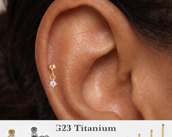 Könnte beinhalten: Ein goldfarbener G23-Titan-Knorpelstecker mit einer kleinen Kugel, an der ein hängender, klarer Zirkonia-Anhänger befestigt ist. Das Set enthält verschiedene Stablängen von 5mm, 6mm, 8mm und 10mm sowie Stärken von 16g, 18g und 20g.