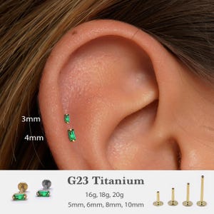 Puede incluir: Primer plano de una oreja con dos pendientes pequeños, rectangulares, de color verde esmeralda, de 3 mm y 4 mm de largo. El texto "G23 Titanium" y las tallas 16g, 18g, 20g, 5mm, 6mm, 8mm y 10mm se muestran.
