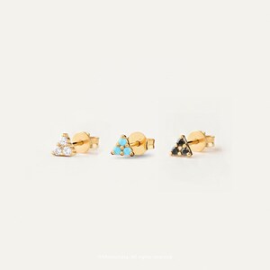 Op de afbeelding: Drie gouden oorknopjes, elk met een driehoekige cluster van drie kleine steentjes. Het eerste oorknopje heeft heldere witte stenen, het tweede heeft fel turquoise stenen en het derde heeft donkere zwarte stenen, allemaal op gouden stiftjes.