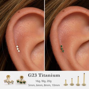 Könnte beinhalten: Goldfarbene G23-Titan-Ohrpiercings mit jeweils drei klaren oder grünen Edelsteinen. Das Bild zeigt die Piercings in einem Ohr. Produktdetails: 16g, 18g, 20g, 5mm, 6mm, 8mm und 10mm.