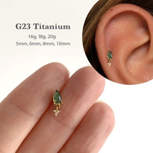 May include: A G23 titanium earring featuring a teardrop-shaped green stone and a small dangling charm with three tiny clear crystals. Text on the image specifies G23 Titanium, 16g, 18g, 20g, and lengths of 5mm, 6mm, 8mm, and 10mm for the jewellery piece.