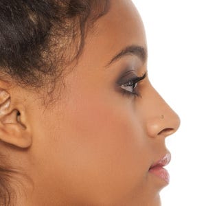 May include: Close-up of a person's face with a small nose ring stud. The person has dark brown hair and dark brown eyes with makeup.