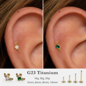 May include: Gold-colored titanium ear studs with a geometric design. One stud features a clear, round gemstone, and the other has a green gemstone. The image also shows the sizes available: 5mm, 6mm, 8mm, and 10mm.