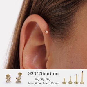 Pode incluir: Um brinco de titânio dourado com um design de estrela, apresentando uma pequena gema transparente no centro. A imagem também mostra o brinco em diferentes tamanhos e comprimentos, com o texto "G23 Titanium" e especificações de tamanho.