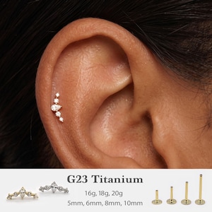May include: A G23 titanium flat back stud earring featuring a curved cluster of clear stones in various sizes, including a pear-shaped centre. The text specifies 16g, 18g, 20g gauges and 5mm, 6mm, 8mm, 10mm lengths for the gold-toned metal posts.