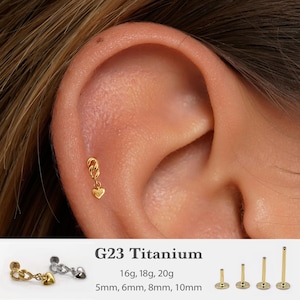 Op de afbeelding: Een gouden titanium oorbel met een kettingontwerp en een hartvormige bedel, getoond op een oor. De afbeelding toont ook andere oorbelstijlen en -maten. Tekst geeft G23 Titanium en maten van 16g tot 20g aan.