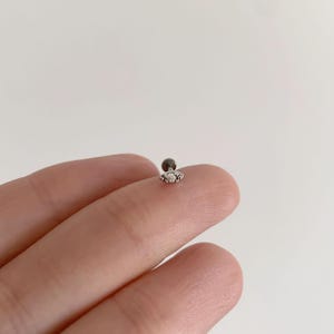 Pode incluir: Um pequeno brinco prateado com uma parte traseira plana e uma frente decorativa com tr&ecirc;s pedras preciosas redondas e transparentes. O brinco &eacute; projetado para um piercing na cartilagem.