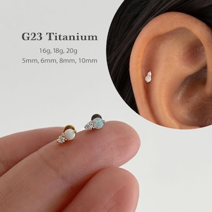 Könnte beinhalten: G23 Titan-Ohrstecker mit einem runden Opal und einem kleineren, klaren Zirkonia-Akzent. Erhältlich in den Stärken 16g, 18g, 20g und Längen 5mm, 6mm, 8mm, 10mm. Der Schmuck ist in Gold- und Silbertönen an einem Ohrknorpel abgebildet.