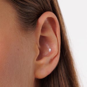 Pode incluir: Close-up de uma orelha com um pequeno brinco oval prateado. O brinco &eacute; cravejado com uma pedra transparente e brilhante. O brinco est&aacute; posicionado na cartilagem interna da orelha.