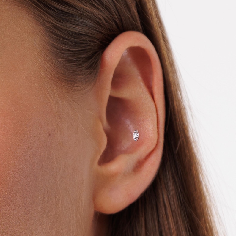 Pode incluir: Close-up de uma orelha com um pequeno brinco oval prateado. O brinco &eacute; cravejado com uma pedra transparente e brilhante. O brinco est&aacute; posicionado na cartilagem interna da orelha.