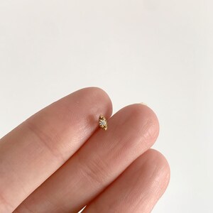 Pode incluir: Um pequeno brinco dourado com uma &uacute;nica pedra preciosa transparente. O brinco tem uma forma curva de crescente. O brinco &eacute; segurado entre dois dedos contra um fundo branco. Uma joia delicada.