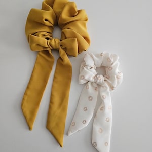 May include: Two hair scrunchies with matching fabric bows. One is a solid mustard yellow and the other is white with a gold floral pattern.