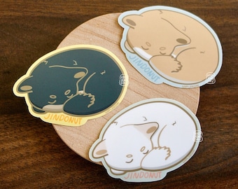 May include: Three round stickers with a cartoon illustration of a sleeping animal on each. The stickers are on a wooden surface. The animals are a brown bear, a black bear, and a white bear. Each sticker has the text "Jinddonut" on it.