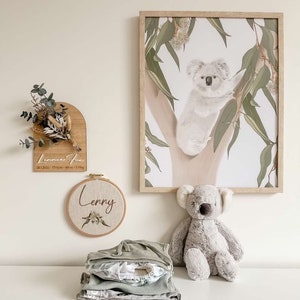 May include: A nursery room decorated with a koala print, a wooden sign with the text "Lennox Fox 28.12.2021 - 7:09pm - 4.6kg - 3.5kg", a round embroidery hoop with the text "Lenny", a gray koala stuffed animal, and a stack of green and white baby clothes.