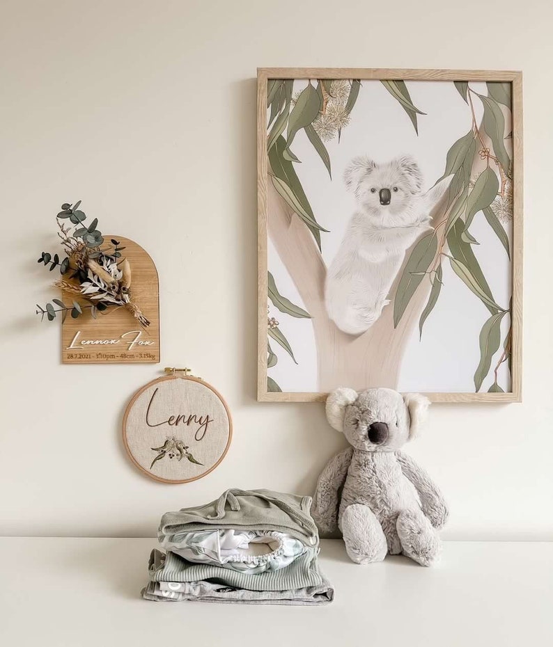 May include: A nursery room decorated with a koala print, a wooden sign with the text "Lennox Fox 28.12.2021 - 7:09pm - 4.6kg - 3.5kg", a round embroidery hoop with the text "Lenny", a gray koala stuffed animal, and a stack of green and white baby clothes.