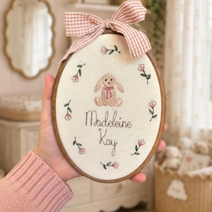 May include: An oval wooden embroidery hoop containing off-white fabric with a hand-stitched design of a tan bunny, pink flowers, and the name Madeleine Kay in brown thread, topped with a decorative pink and white gingham fabric bow.