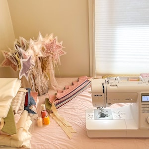 May include: A white sewing machine with a pink table top. There are spools of thread, fabric scraps, and a stack of white fabric on the table. A bouquet of pink and white star-shaped fabric wands are in a vase on the left side of the table.