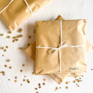 May include: Three brown paper packages tied with white string and labeled with names: Mabel, Kenzie, and Mia. The packages are on a white surface with gold star confetti.