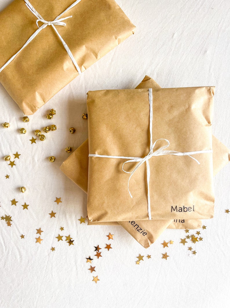 May include: Three brown paper packages tied with white string and labeled with names: Mabel, Kenzie, and Mia. The packages are on a white surface with gold star confetti.