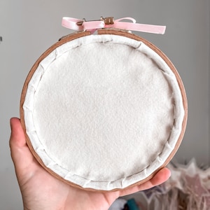 May include: A wooden embroidery hoop with a white fabric stretched across the inside. The hoop is tied with a pink ribbon.
