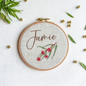 May include: A white embroidery hoop with a brown wooden frame. The hoop features a floral design with red flowers and green leaves. The name "Jamie" is embroidered in brown cursive lettering.