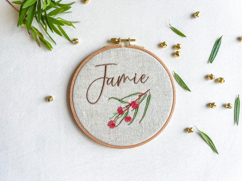 May include: A white embroidery hoop with a brown wooden frame. The hoop features a floral design with red flowers and green leaves. The name "Jamie" is embroidered in brown cursive lettering.